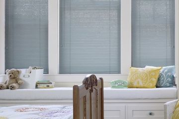 a window seat with pillows and a teddy bear, covered by light-colored aluminum blinds.