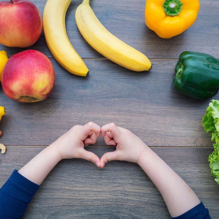 A kid making a heart with their hands surrounded by fruit and vegetables.