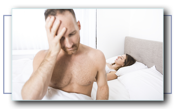 Man sitting on his bed with his head in his hands, while a woman sleeps in the bed in the background.