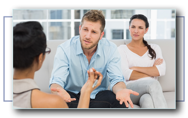 Unhappy white couple sitting across from a couples counselor. The man is talking to the counselor concerned, while the woman is sitting back with her arms crossed.