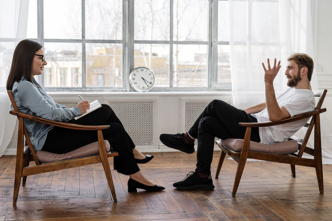 A Mental Health Therapist Facing a Talking Patient as a Clock Ticks Between them on a Windowsill