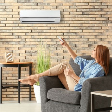 woman on easy chair pointing remote at mini split on brick wall, smiling