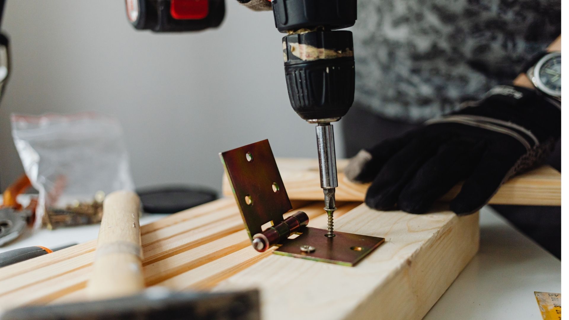 A close-up action shot of a handyman in black work gloves using a power drill to install a brass hinge onto a wood plank
