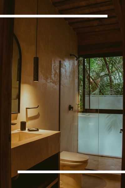 A spa-inspired bathroom with warm, ambient lighting, featuring a smooth concrete or plaster vanity, a modern black pendant light, and a view of tropical greenery through a frosted glass door.
