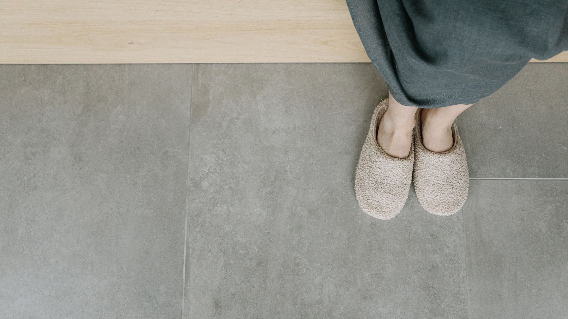 An overhead minimalist shot of a person wearing cozy beige slippers standing on a newly installed large-format gray stone-look tile floor