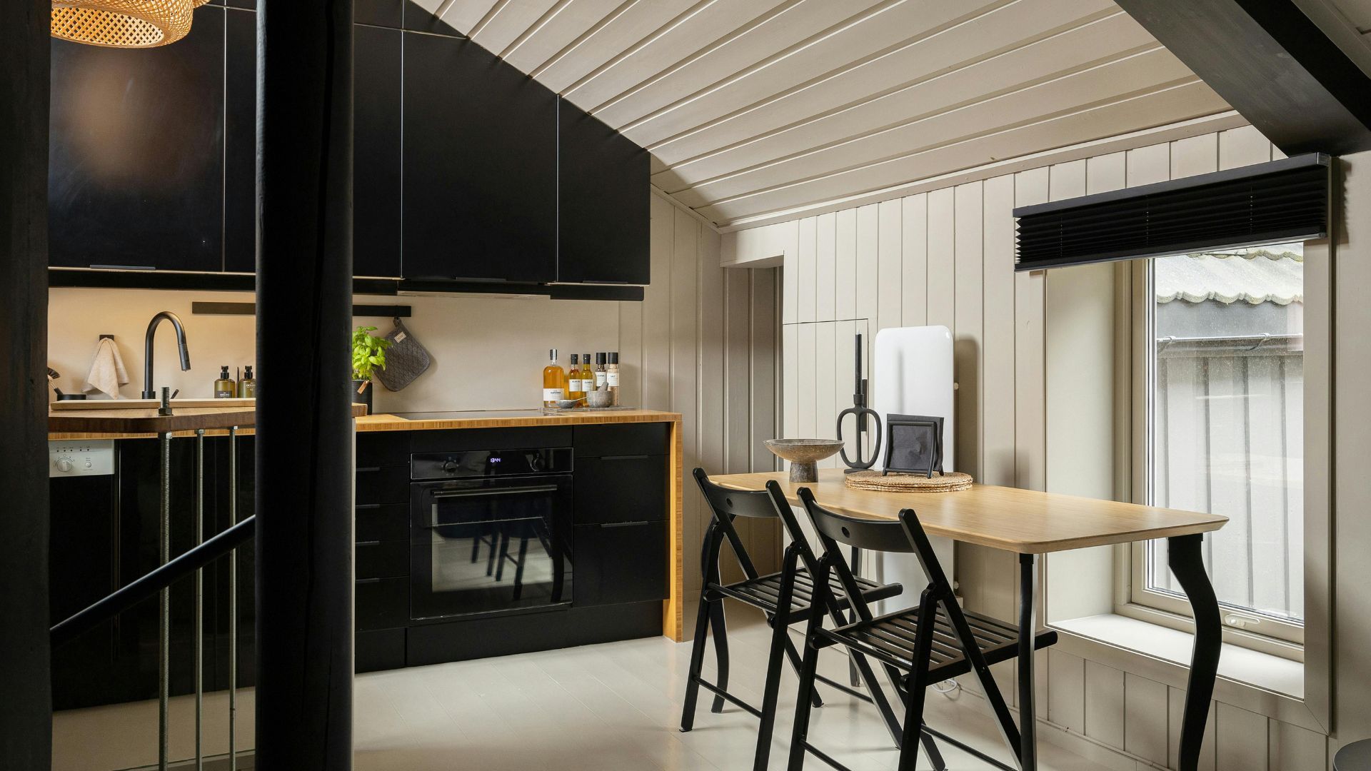 A stylish, compact modern kitchen and dining nook featuring matte black cabinetry, light wood countertops, and a minimalist black folding chair set against vertical off-white shiplap walls