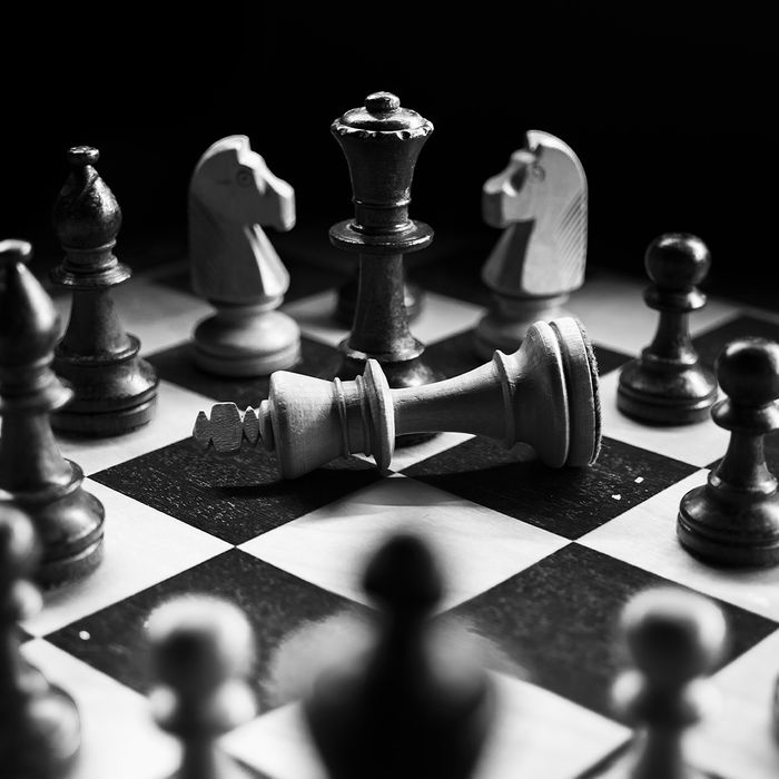 Black and white picture of a chessboard with the white king laying on its side and all other pieces surrounding it