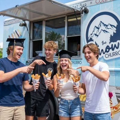 Four laughing teenagers, including two graduates, holding churros in front of 'The Snowy Churro' light blue food truck