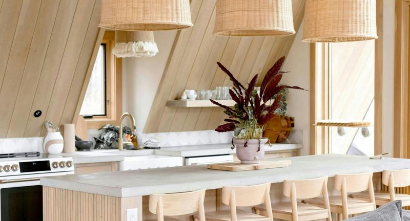 A kitchen island with a thick concrete countertop and light wood fluted paneling. Natural-toned wooden chairs are lined up underneath. Large woven wicker pendant lights hang from a slanted, wood-planked ceiling. A kitchen island with a thick concrete countertop and light wood fluted paneling. Natural-toned wooden chairs are lined up underneath. Large woven wicker pendant lights hang from a slanted, wood-planked ceiling.