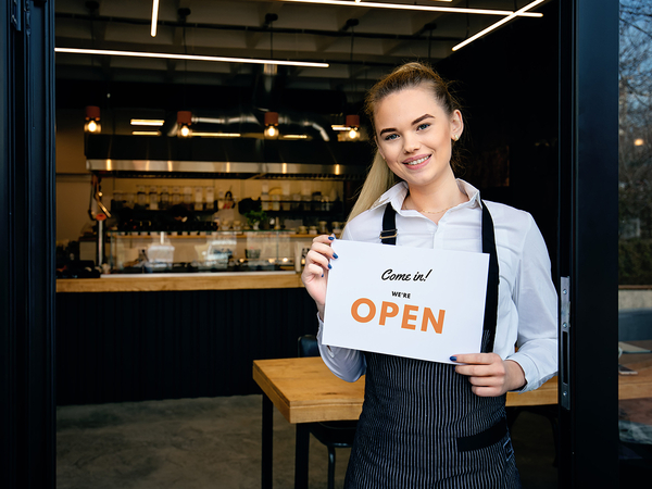 Local business woman standing in front of a coffee shop holding a sign that says “Come in! We’re open.”