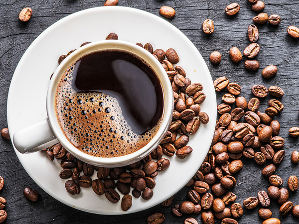 A cup of freshly roasted coffee sitting on a plate, surrounded by different kinds of coffee beans. 