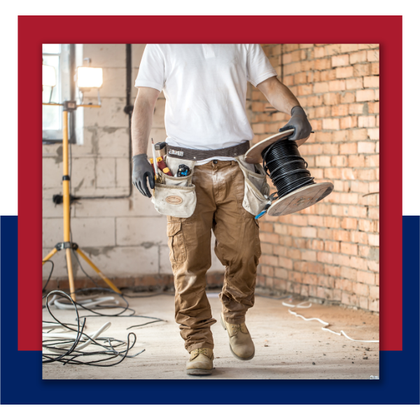A professional in a white shirt and work trousers carrying a large spool of black electrical wire through a residential construction site.