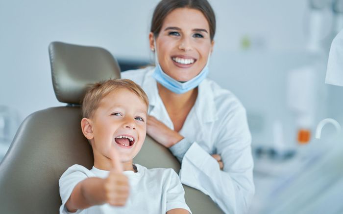 a child in a dental chair giving a thumbs up with a dentist behind them smiling