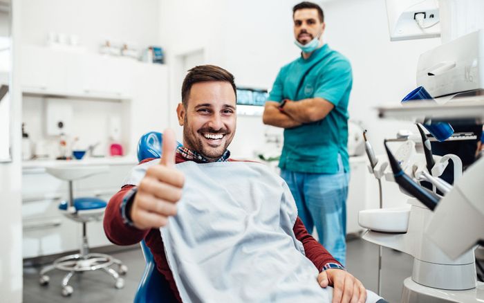 a man in a dental chair giving a thumbs up with a dentist in the background