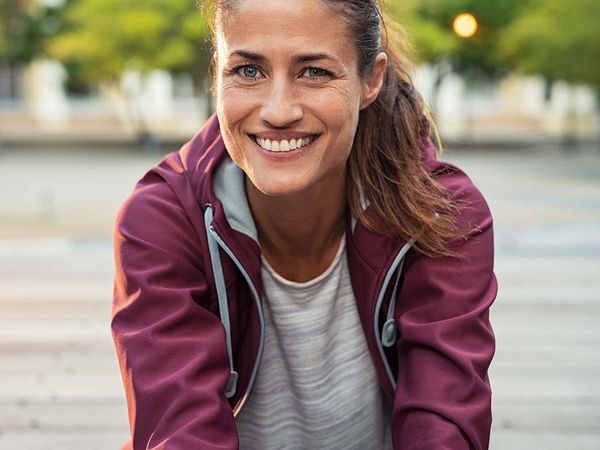 Portrait of smiling woman sitting on floor of city street after running. Healthy mature runner resting after workout exercise and looking at camera. Active latin sporty woman enjoying outdoors in autumn.