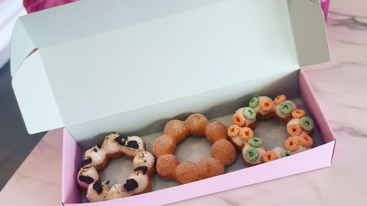 A top-down view of a light pink box containing three Mochi Donuts. From left to right: a donut with white glaze and crushed Oreo chunks, a classic cinnamon sugar-dusted donut, and a white-glazed donut topped with colorful Froot Loops cereal.