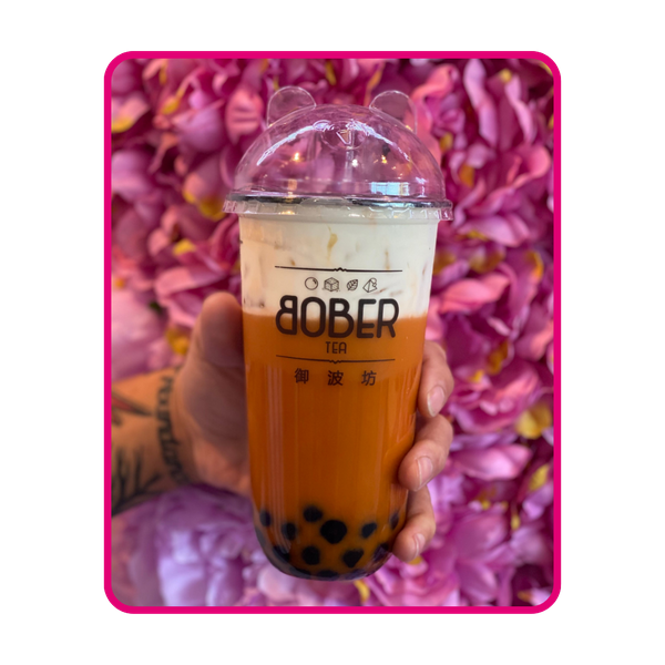 A vibrant, orange-layered Thai Milk Tea held against a floral pink background. The drink shows distinct layers of dark tea and creamy white milk, with chewy boba pearls settled at the bottom and a cute bear-eared lid.