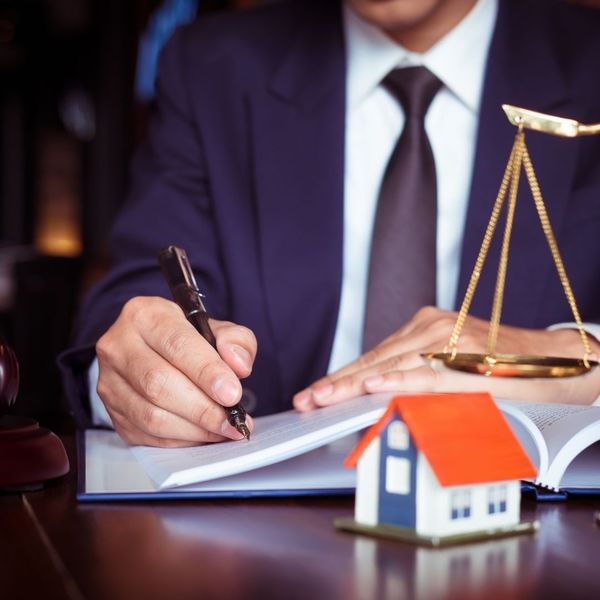 A lawyer in a suit signs documents at a desk with a small model house and a scale of justice nearby.