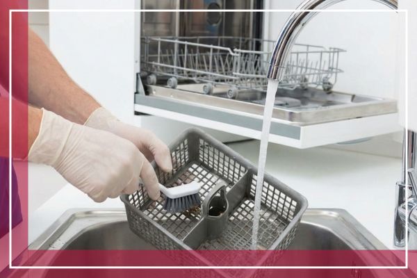A person wearing gloves cleans a dishwasher filter basket with a brush under running water in a sink.