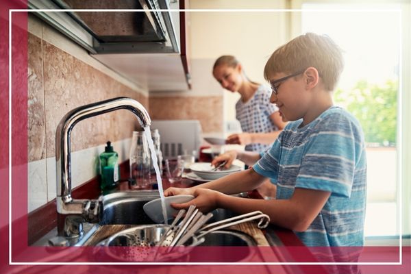 A young boy in a blue and white striped shirt washes dishes in a kitchen sink, while a girl stands in the background Old Westbury 1.jpg