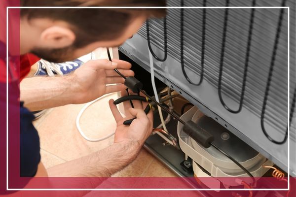 A person's hands are shown using pliers on the wiring at the back of a refrigerator Matinecock 3.jpg