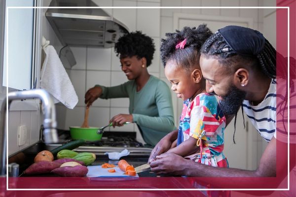 A family in the kitchen. A man is helping a young girl cut vegetables, while a woman stirs a pot on the stove in the background Locust Valley 4.jpg