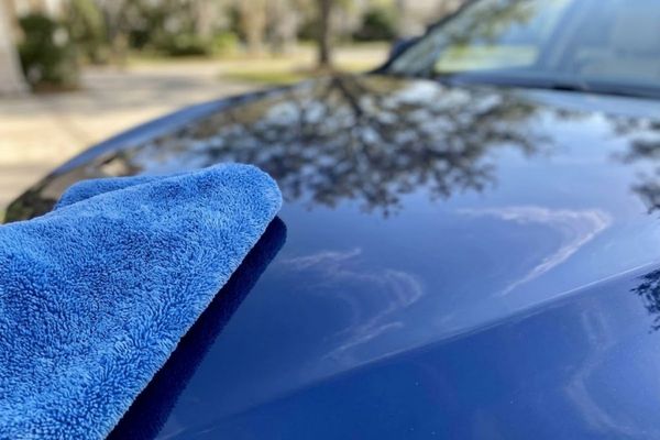 a plush microfiber towel gently wiping down a highly reflective, freshly waxed car hood. The pristine reflection in the car's paint shows a clear blue sky and a subtle tree canopy.