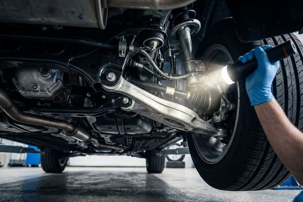a mechanic inspecting the underside of a vehicle's suspension system using a bright mechanic's flashlight. Focus is on the clean mechanical components, metal textures, and the light beam.