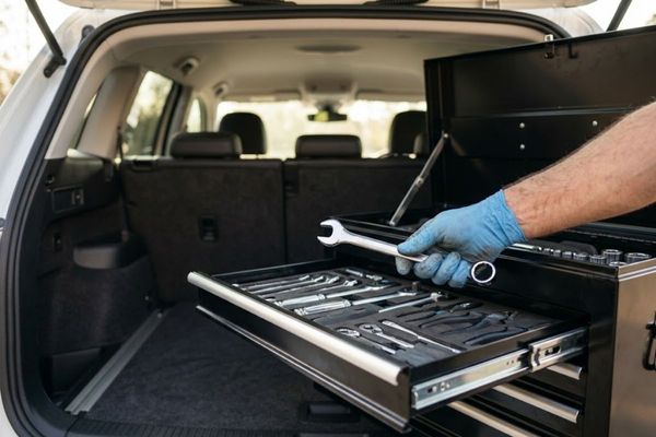a mechanic's toolbox open in the back of a vehicle, with the mechanic's hand reaching for a polished silver wrench. The tools are neatly organized and gleaming under natural daylight.