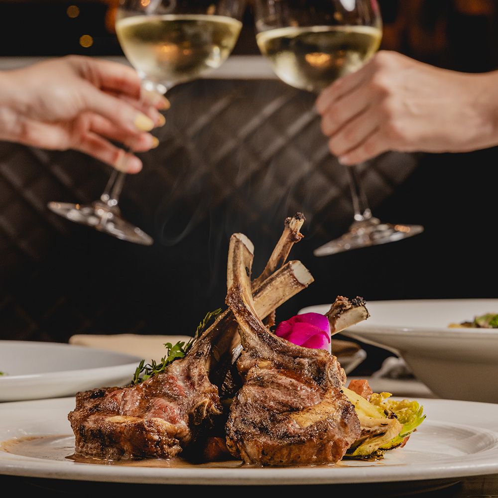 two people doing cheers with wine glasses behind a steak dish