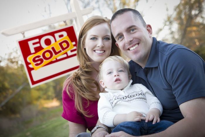 a couple holding their toddler in front of a sold sign