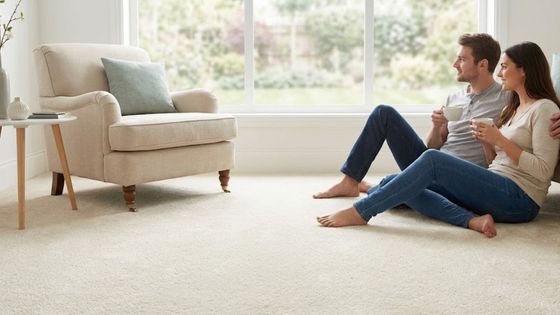 A comfortable couple relaxes on soft cream carpet in a naturally lit living room, enjoying coffee and a view through a large window.
