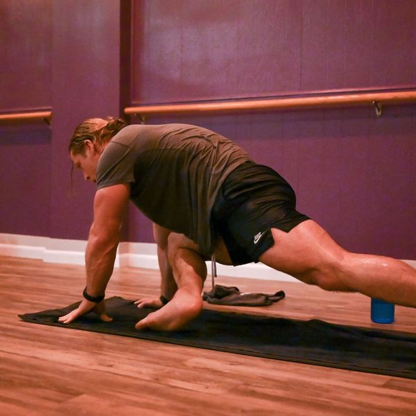 A man in a grey t-shirt and black shorts performing a deep lizard lunge on a black yoga mat in a studio with purple walls.
