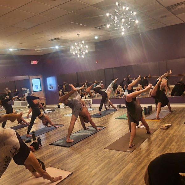 A wide shot of a yoga class in a purple studio with mirrors, where several students are practicing a three-legged downward dog pose.