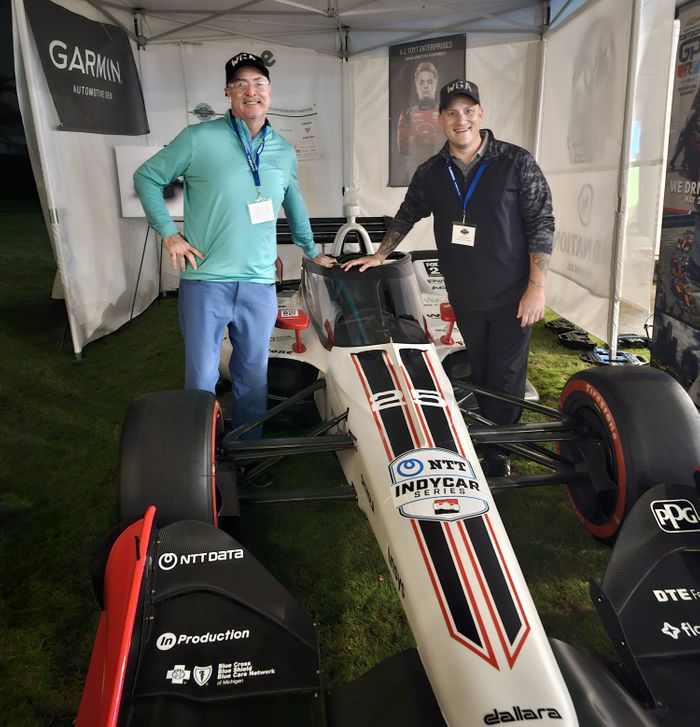 Two men wearing event lanyards and hats smiling next to a white, black, and red NTT IndyCar under a white event tent. A Garmin sign is visible in the background. WGA Representatives with IndyCar at GARMIN Event