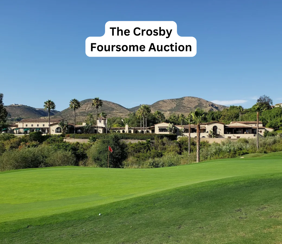 A scenic view of a vibrant green golf course putting green featuring a red pin flag in the foreground. In the background, a sprawling, elegant Spanish-style clubhouse is nestled among palm trees and lush vegetation, set against a backdrop of rolling hills under a clear blue sky. A prominent white text box centered at the top reads "The Crosby Foursome Auction". The Crosby Foursome Auction Golf Course and Clubhouse View