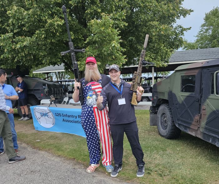 Two men smiling and posing with military rifles next to a parked Humvee at an outdoor event. One man is wearing American flag overalls, and the other is wearing a Warrior Golf Academy hat and event lanyard. A blue 125th Infantry Regiment Association banner is visible in the background. WGA and 125th Infantry Regiment Association Display