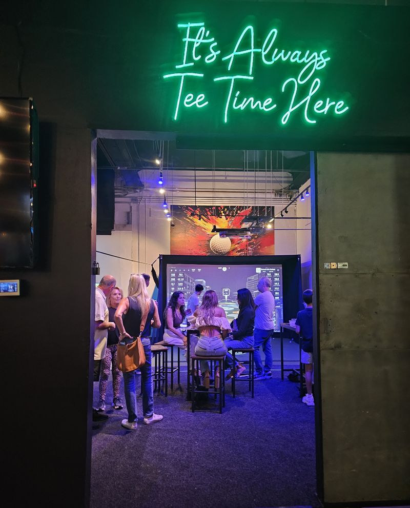 A group of guests gather inside an indoor golf simulator bay at Strokes N Drivers in Fort Lauderdale, under a neon sign that reads "It's Always Tee Time Here" to celebrate the launch of Warrior Golf Academy’s online community for Veterans, First Responders, and supporters. Warrior Golf Academy Community Launch Indoor Golf Event