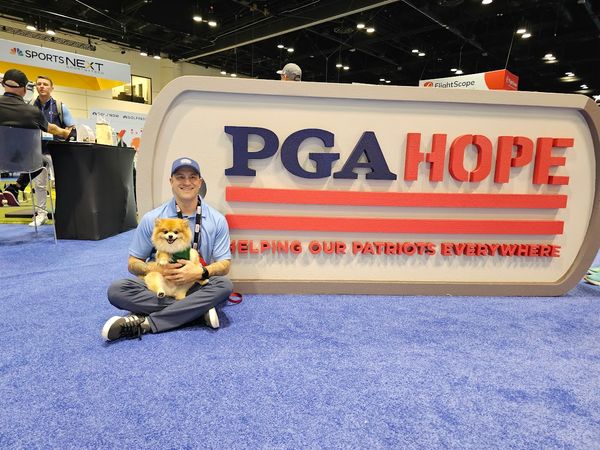 A smiling veteran in a blue polo and cap sits cross-legged on the floor at the PGA HOPE exhibit booth, holding a small service dog. Behind him is a large PGA HOPE sign with the slogan “Helping Our Patriots Everywhere,” displayed on a blue carpeted floor at a golf industry event. The setting promotes veteran support through golf, community, and wellness initiatives. Jonathan Sessa Army Military Veteran at PGA HOPE Booth with Service Dog