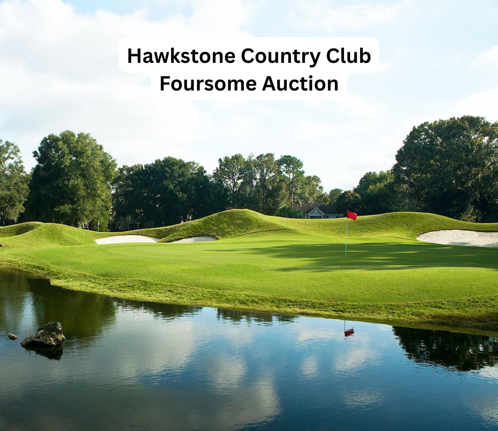A picturesque view of a lush green golf course putting green at Hawkstone Country Club. The green features dramatic, rolling grass mounds and hills in the background, bordered by pristine white sand bunkers and a dense line of mature trees. A vibrant red pin flag stands on the green, beautifully reflecting alongside the blue sky and fluffy white clouds in the calm water hazard in the foreground. A small rock rests near the water's edge on the left. A rounded white text box centered at the top reads "Hawkstone Country Club Foursome Auction". Hawkstone Country Club Foursome Auction Green and Pond View