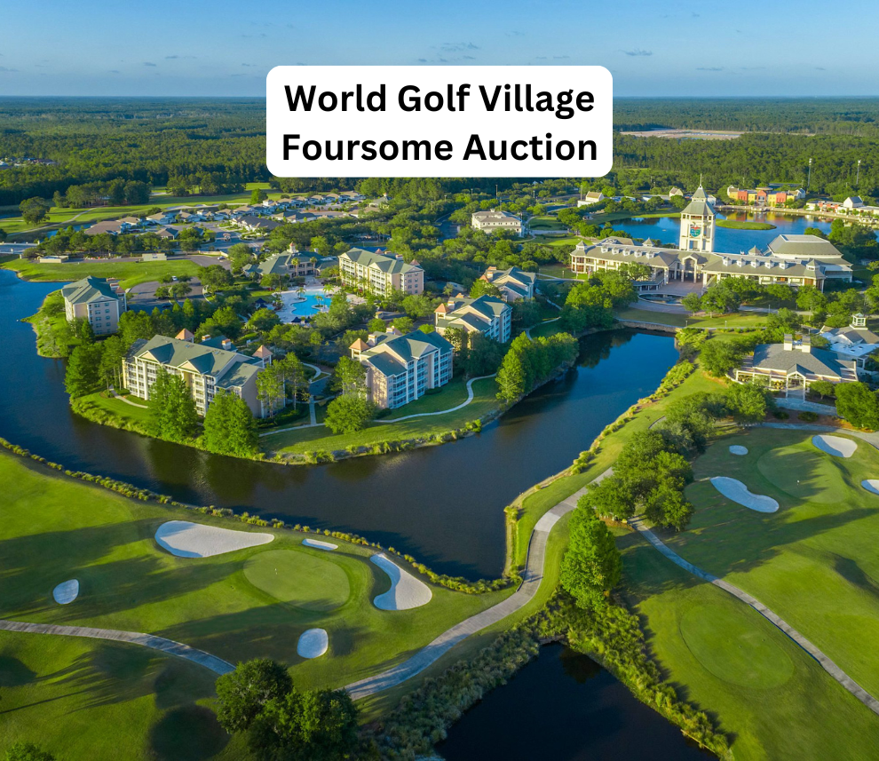 An expansive, high-angle aerial view of World Golf Village under clear daylight. In the foreground, beautifully manicured green golf course fairways and sand bunkers are visible alongside winding water hazards. Across the water sits a resort community with multi-story hotel buildings, a swimming pool, and the prominent World Golf Hall of Fame clubhouse and tower structure. A vast forest line extends across the horizon under a soft blue sky. A rounded white text box centered at the top reads "World Golf Village Foursome Auction". World Golf Village Foursome Auction Aerial View
