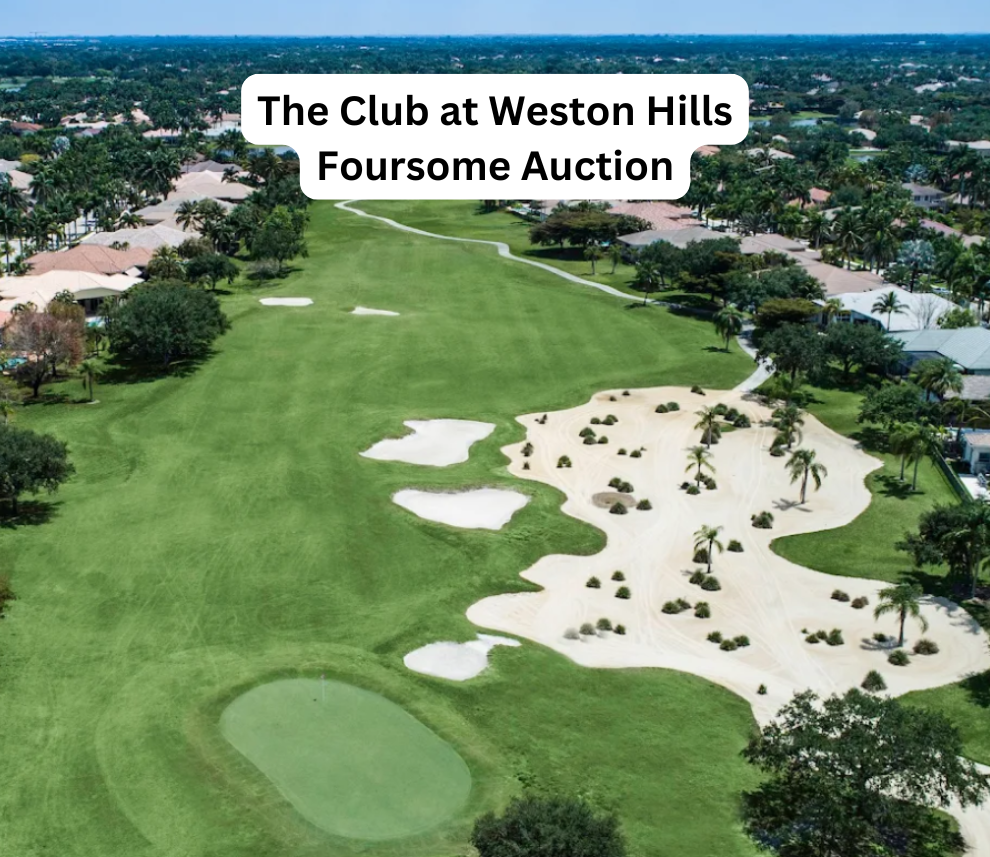 An aerial drone view looking down a long, vibrant green golf course fairway at The Club at Weston Hills. A green putting green sits in the lower left foreground, bordered by several white sand bunkers. To the right, a massive, uniquely shaped waste bunker is filled with white sand, small desert shrubs, and several palm trees. Winding golf cart paths and residential homes line both sides of the expansive course under a clear blue sky. A rounded white text box centered at the top reads "The Club at Weston Hills Foursome Auction". The Club at Weston Hills Foursome Auction Aerial View