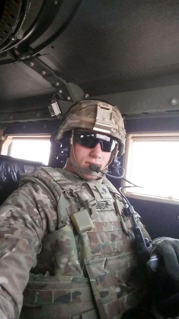 A U.S. Army soldier in full combat gear, including helmet, ballistic vest, sunglasses, and headset, takes a selfie inside a military vehicle. The name tag "SESSA" is visible on the vest. The image captures the readiness and professionalism of a service member during deployment or training operations. Army Soldier Jonathan Sessa in Tactical Gear Inside Military Vehicle