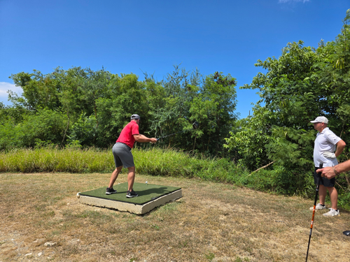 A landscape photograph from our deployment to Naval Station Guantanamo Bay, Cuba, showing a golfer in a red shirt and grey shorts standing on an artificial turf mat atop a concrete block tee box. He is in the middle of his backswing, preparing to drive a golf ball into the rugged, un-irrigated environment. Another player in a white shirt and hat stands to the right watching the shot, surrounded by dense tropical brush and arid, dry grass. The image highlights the extreme, raw conditions of the course, emphasizing Warrior Golf Academy’s mission to bring high-energy relief, over $30,000 in tournament prizes, and brand-new equipment to our forward-deployed Troops as part of the USA 250th Secured Horizon Series. Service Member Preparing to Tee Off From Concrete and Turf Tee Box at Naval Station Guantanamo Bay GTMO Golf Tournament