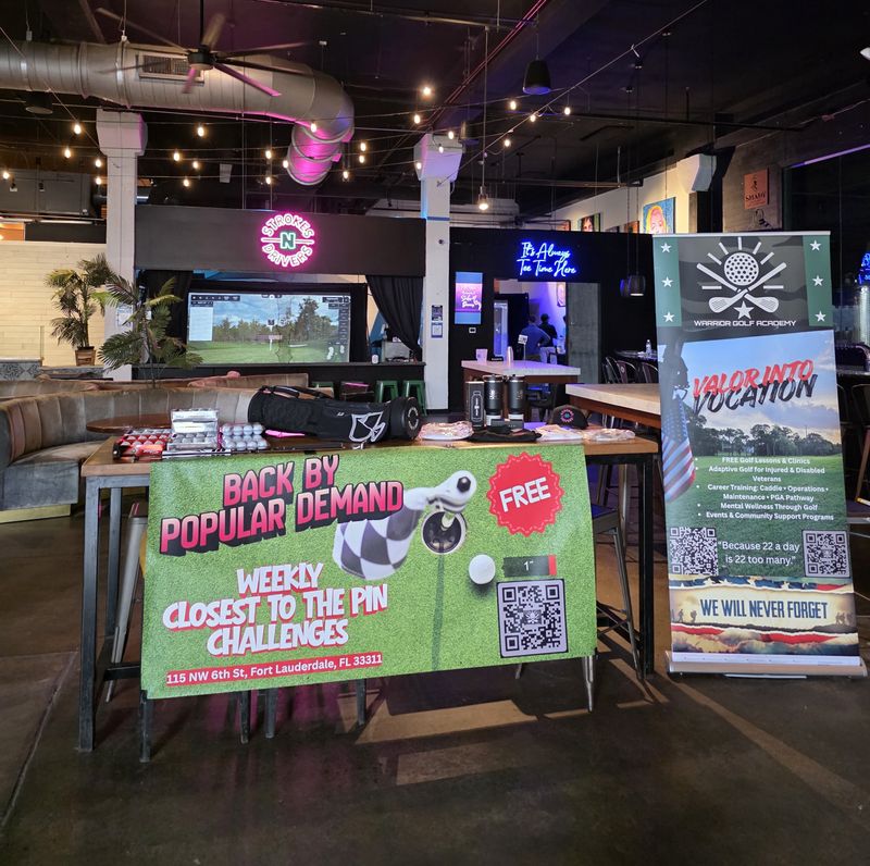 Interior view of Strokes N Drivers in Fort Lauderdale featuring the Warrior Golf Academy 'Valor Into Vocation' partnership display. The image shows promotional banners for weekly 'Closest to the Pin' challenges and information on free golf clinics, adaptive golf for injured veterans, and career training programs. This partnership highlights how Strokes N Drivers serves as a community hub for South Florida Veterans and First Responders, combining high-tech golf entertainment with a mission-first approach to support mental wellness and peer connection for those who served. Warrior Golf Academy Valor Into Vocation Display at Strokes N Drivers Fort Lauderdale