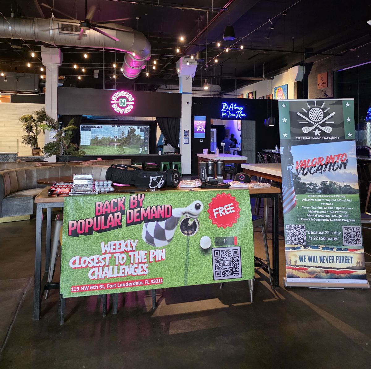 Interior view of Strokes N Drivers in Fort Lauderdale featuring the Warrior Golf Academy 'Valor Into Vocation' partnership display. The image shows promotional banners for weekly 'Closest to the Pin' challenges and information on free golf clinics, adaptive golf for injured veterans, and career training programs. This partnership highlights how Strokes N Drivers serves as a community hub for South Florida Veterans and First Responders, combining high-tech golf entertainment with a mission-first approach to support mental wellness and peer connection for those who served. Warrior Golf Academy Valor Into Vocation Display at Strokes N Drivers Fort Lauderdale