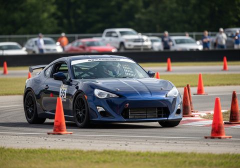 A blue sports car, labeled with the number 55, skillfully maneuvers through a course marked by orange cones on a racetrack. The driver is visible through the windshield, wearing a helmet, and the car is surrounded by a green grassy area and a blurred background with spectators and other vehicles. Blue Race Car Navigating Cones on Racetrack