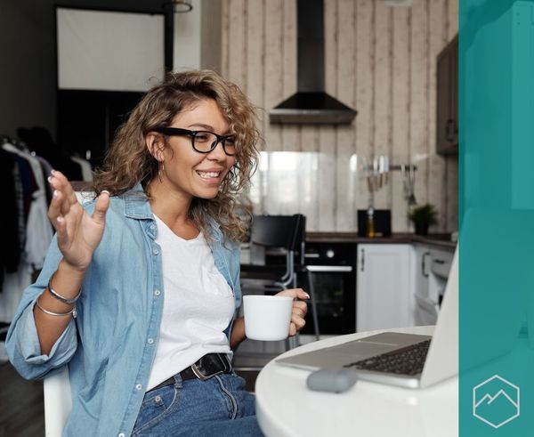 A woman in a kitchen smiling and waving at a laptop screen to telehealth provider. A woman in a kitchen smiling and waving at a laptop screen to telehealth provider.