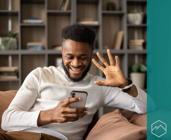 A man laughing and waving at his phone while sitting on a couch on a telehealth video call. A man laughing and waving at his phone while sitting on a couch on a telehealth video call.