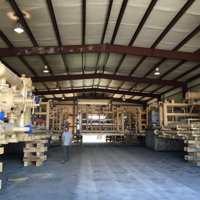 A wide interior shot of a large industrial warehouse, housing multiple oversized pieces of beige oil and gas machinery on wooden blocks, with a person standing in the center of the spacious concrete floor.